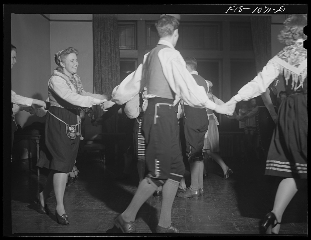 Seven dancers in 1940s dress form a circle holding hands indoors, a Swedish folk dance in progress.