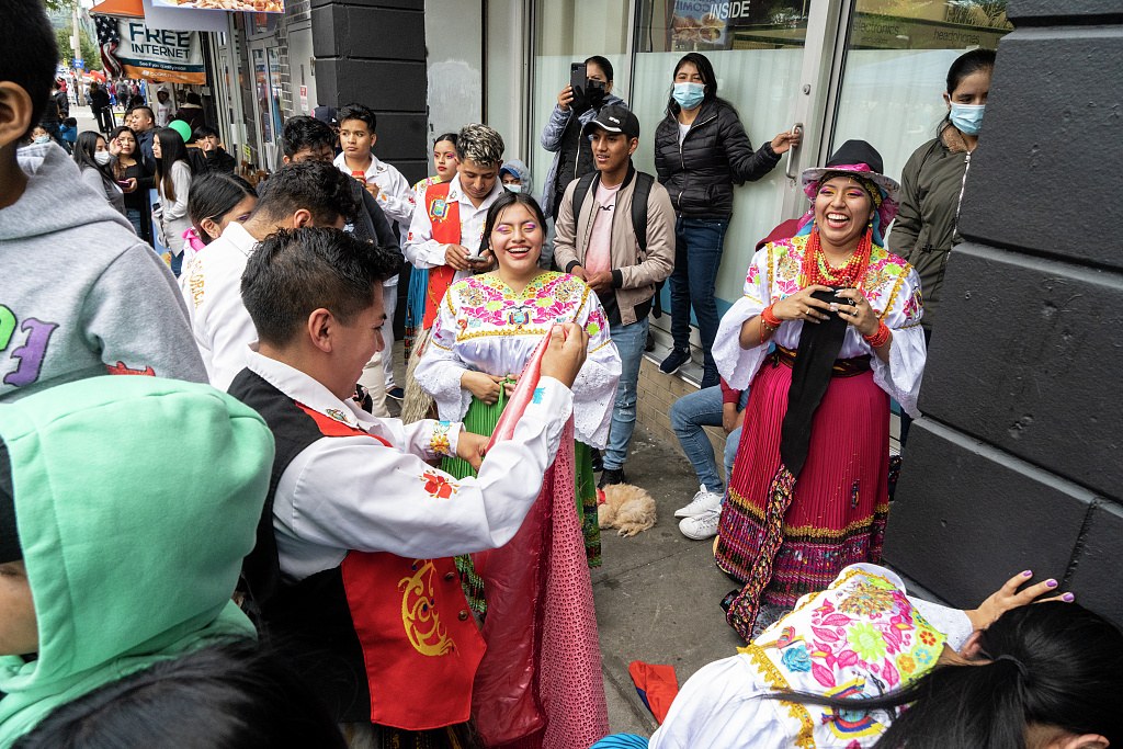 Two women in embroidered Ecuadorian traditional dress stand smiling among a crowd on a Queens street corner, resting between dances.