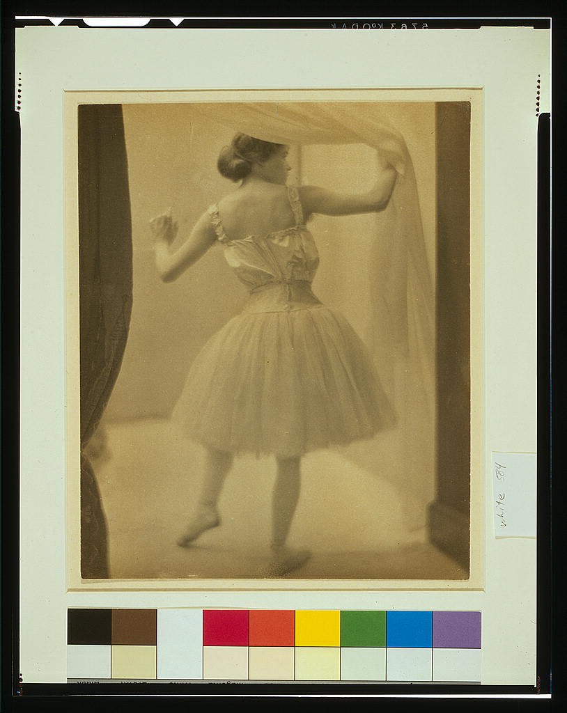 Young woman in tutu and fitted bodice stands in profile with raised arm, demonstrating classical ballet pose in studio setting.