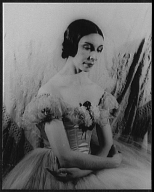 Studio portrait of a young woman in classical ballet costume: fitted bodice, short sleeves, full tulle skirt. Dark hair styled in 1940s waves, standing upright