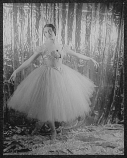 Young woman in classical ballet tutu and bodice stands centered against draped backdrop, arms extended at shoulder height, in formal studio portrait.