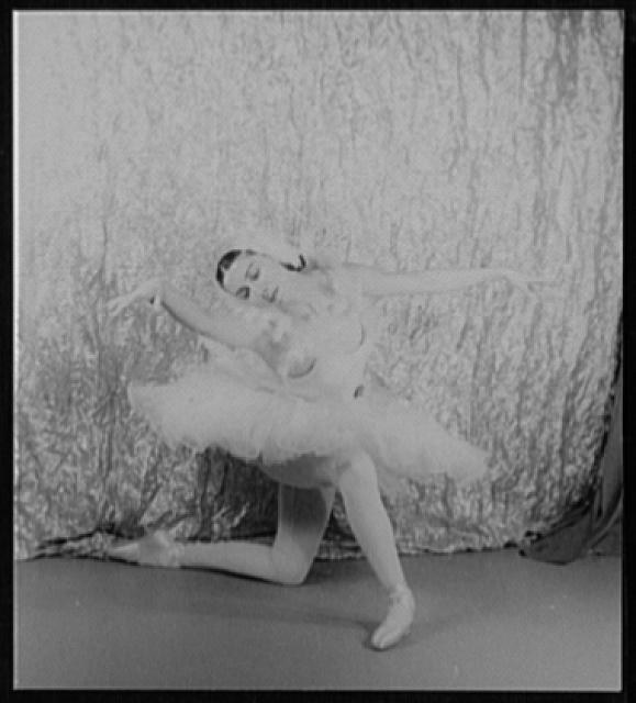 Alicia Markova in ballet tutu, kneeling and arched backward with head tilted back, against dark curtains.