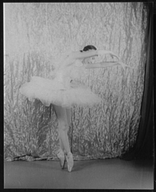 Black-and-white photograph of a female dancer in a white tutu on pointe, her body tilted forward with arms extended, against a textured backdrop.