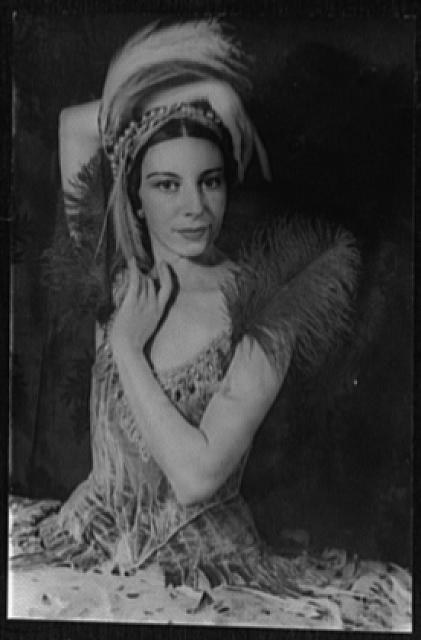 Black-and-white photograph of a woman in elaborate white headdress and embroidered bodice, arms gracefully posed, seated against a dark background.