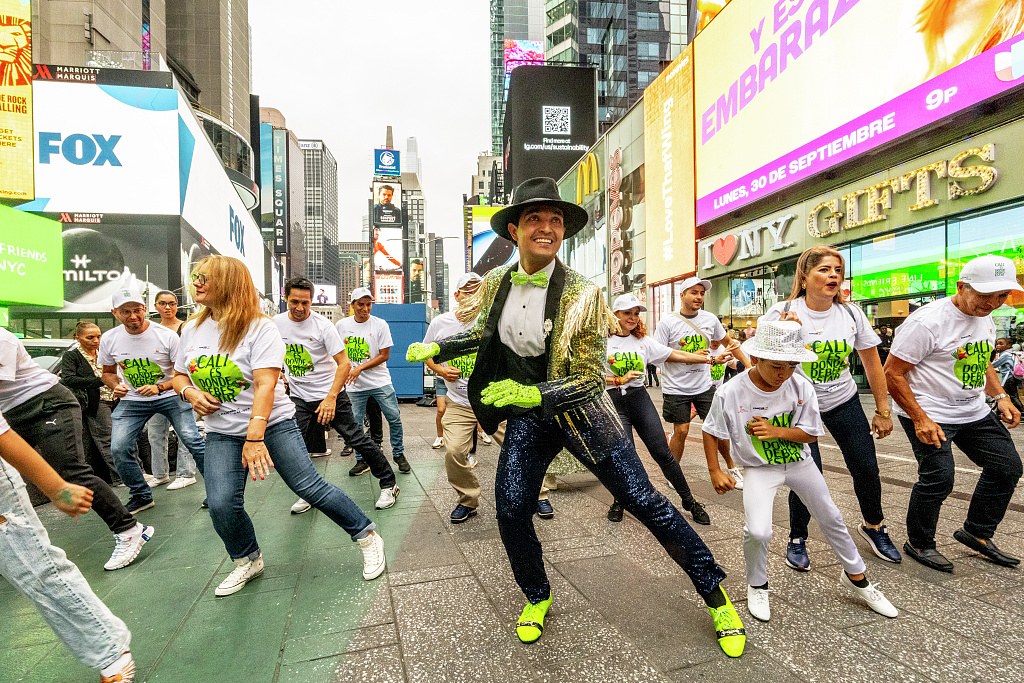 A man in gold jacket and neon shoes leads a crowd of white-shirted dancers in a street salsa celebration in Times Square.