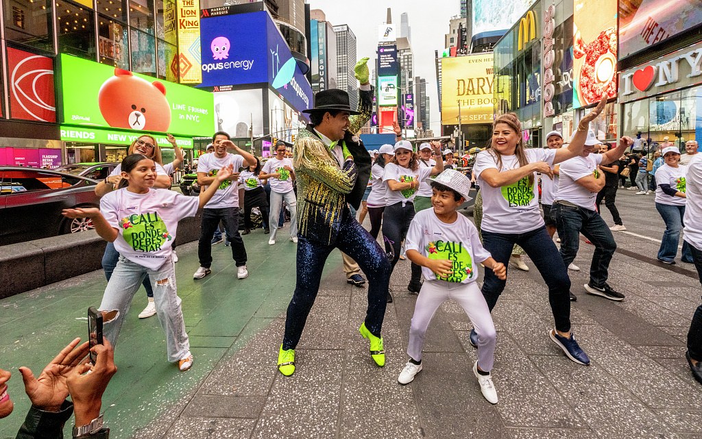 Large group of salsa dancers in white t-shirts dance in Times Square with a costumed salsero in gold lamé and neon accents at center.