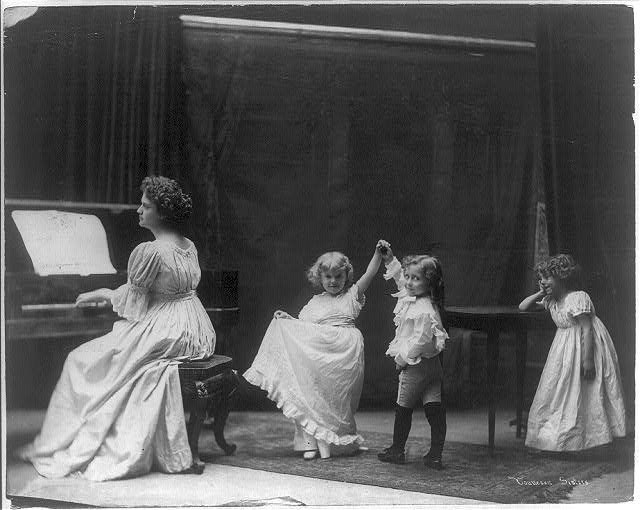 A woman at a piano accompanies three children in white dresses learning to dance in an Edwardian parlor.