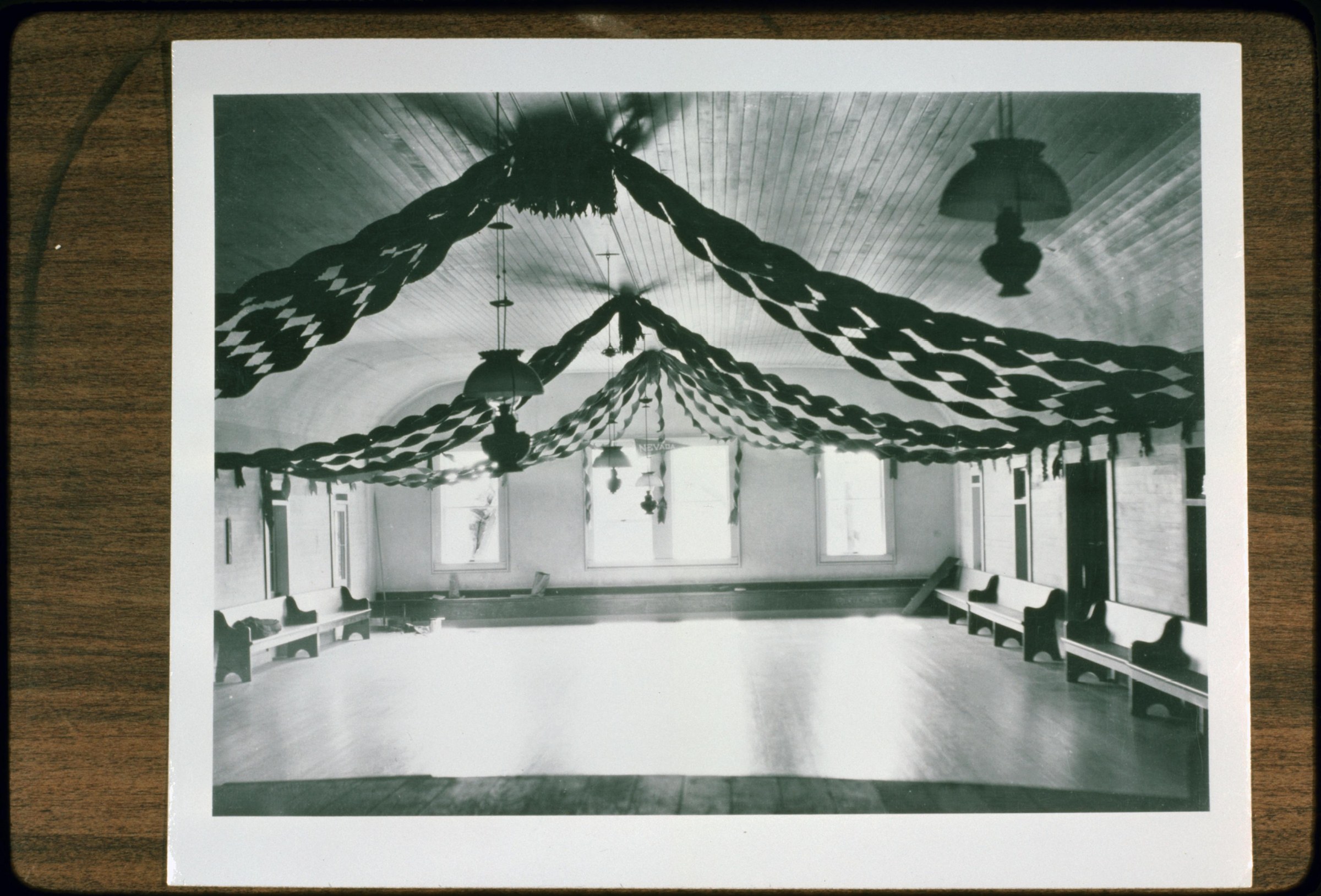 Empty wood-floored dance hall decorated with swags and garland, bright windows along walls, wooden benches for seating.