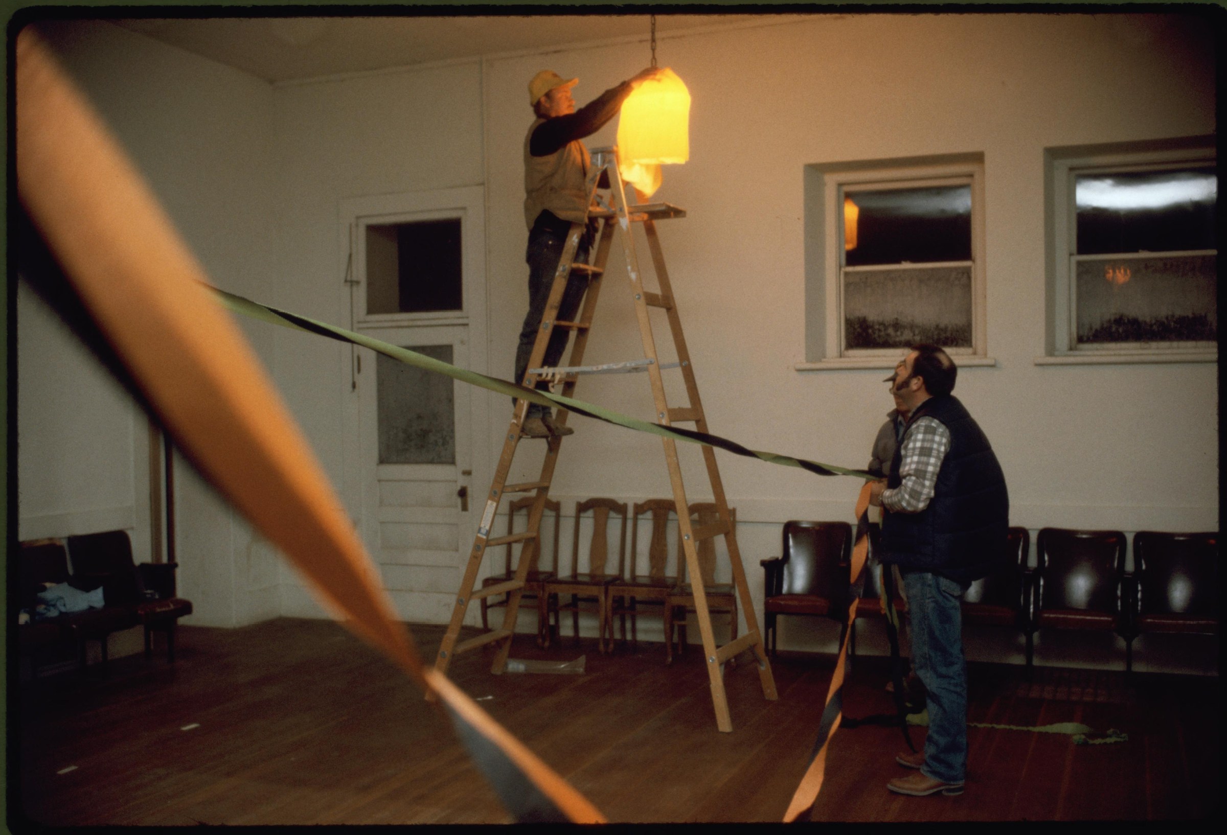 Two men work together to install a bright yellow pendant lamp in a wooden-floored hall with stacked chairs along the wall.