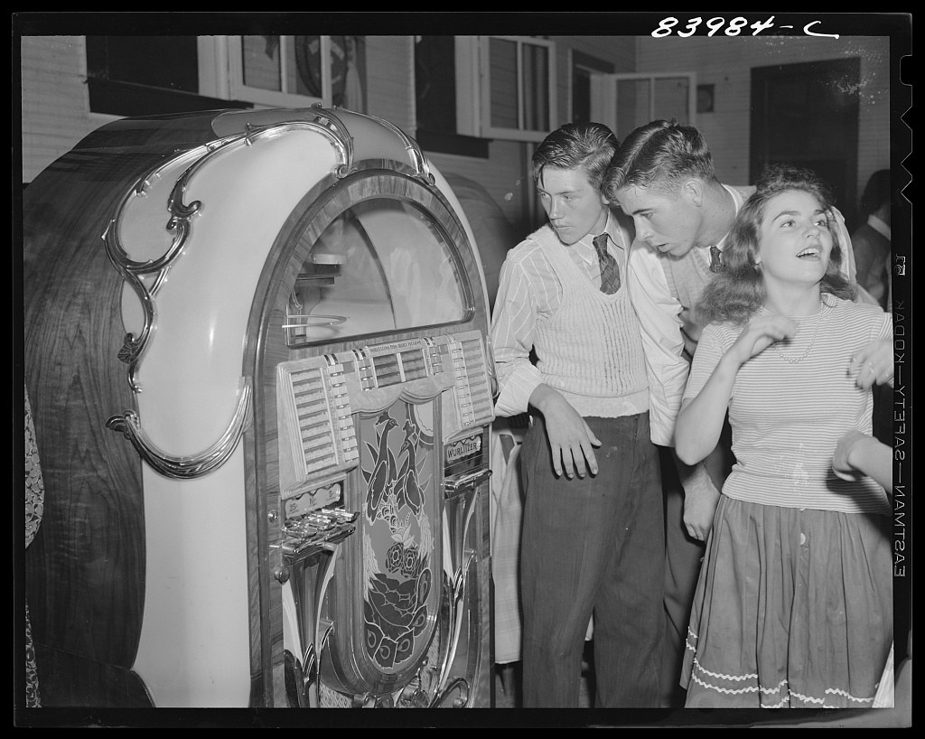 Three young people stand before an open jukebox in a dimly lit dance hall, gazing at the machine with interest.