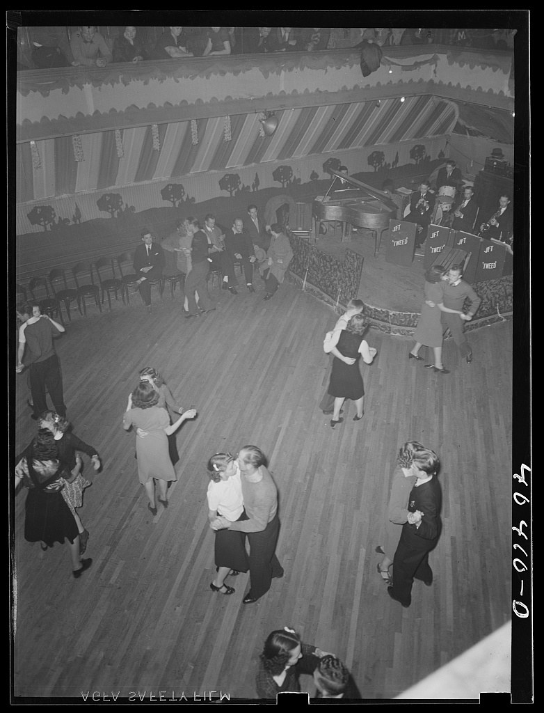 Overhead view of couples dancing in closed ballroom hold on a wooden dance floor, with an orchestra platform and piano visible in a 1930s-era decorated dance
