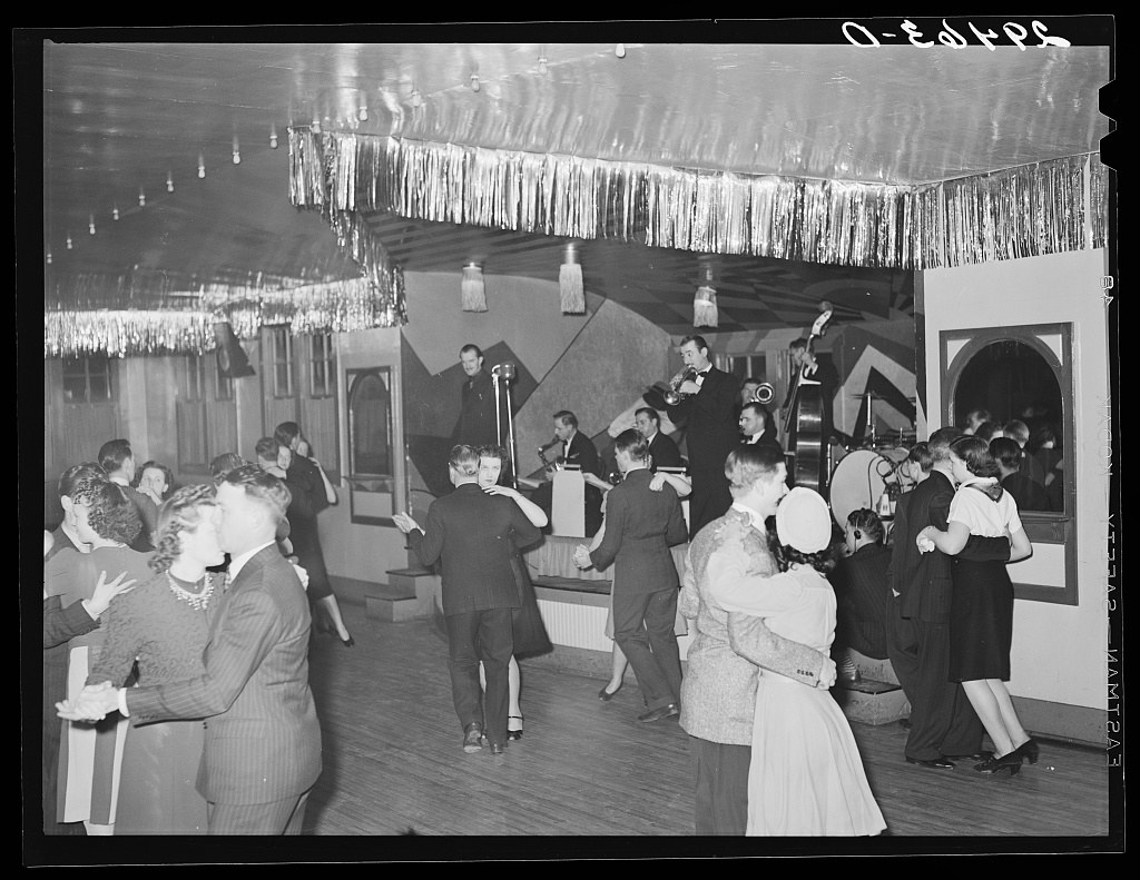 Crowded black-and-white photograph of a 1940s ballroom with couples in formal dress dancing to live music in closed ballroom holds.