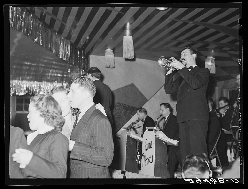 Black-and-white photograph of couples dancing in a decorated hall; a man in a suit and woman in a dark dress dance in the foreground while a trumpet player and