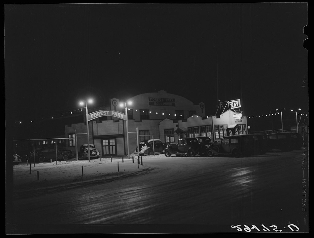 Night photograph of Forest Park dance hall in Marshalltown, Iowa, lit by floodlights and strings of lights, with 1940s automobiles in the parking lot.