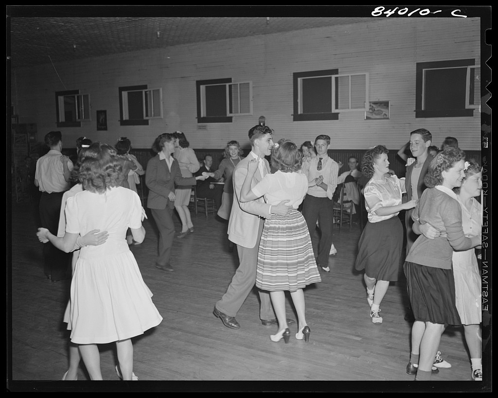 About thirty dancers in couples and small groups fill a brick dance hall, men in suits and military uniforms, women in simple 1940s dresses, dancing informally.