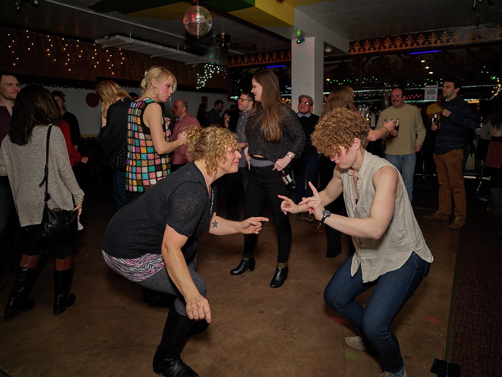 Two women in casual dress squat facing each other with arms extended in a playful dance pose at an indoor community celebration.