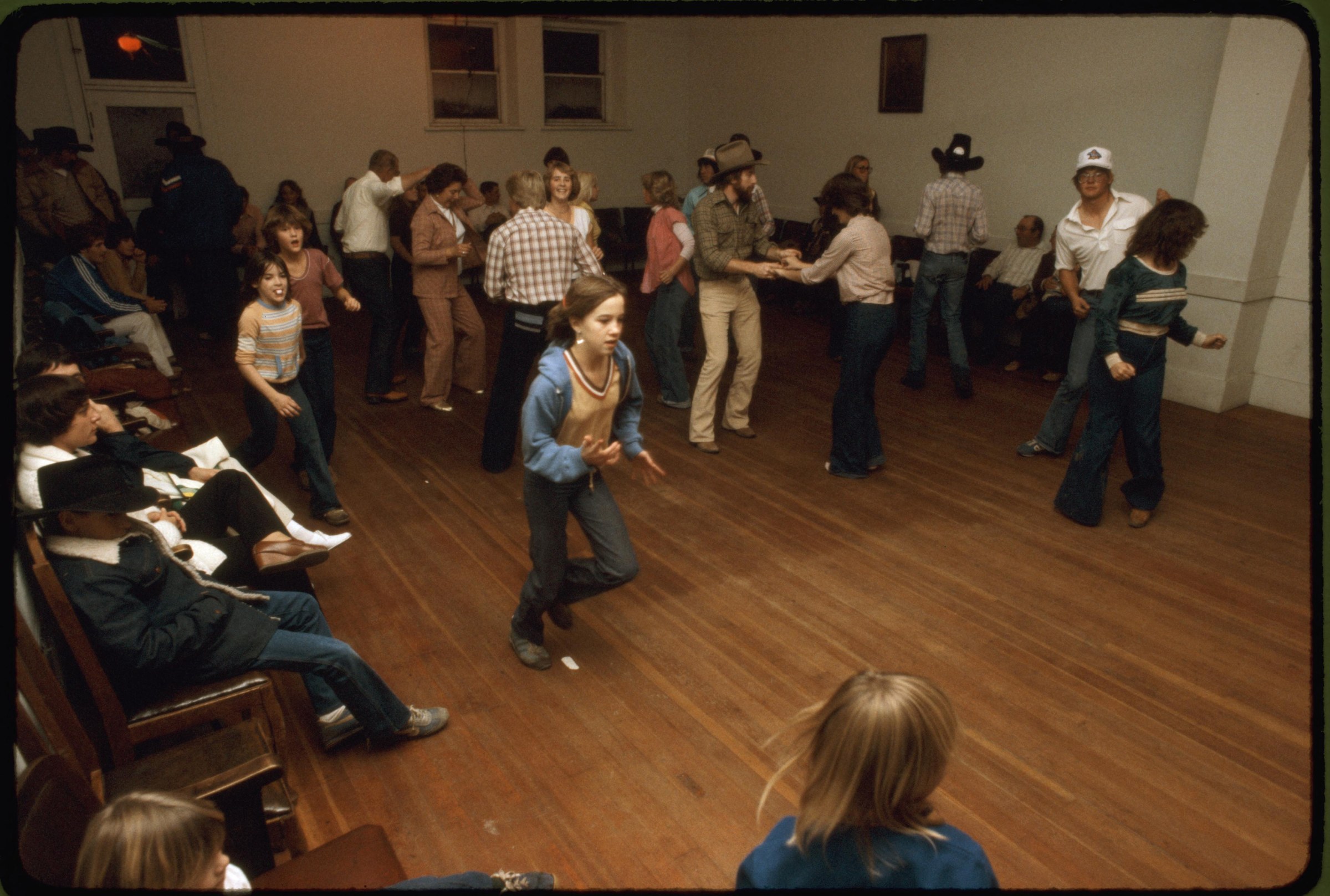 Large group of adults and children in casual 1970s dress stand and dance in a gymnasium, some wearing cowboy hats, gathered for a community event.