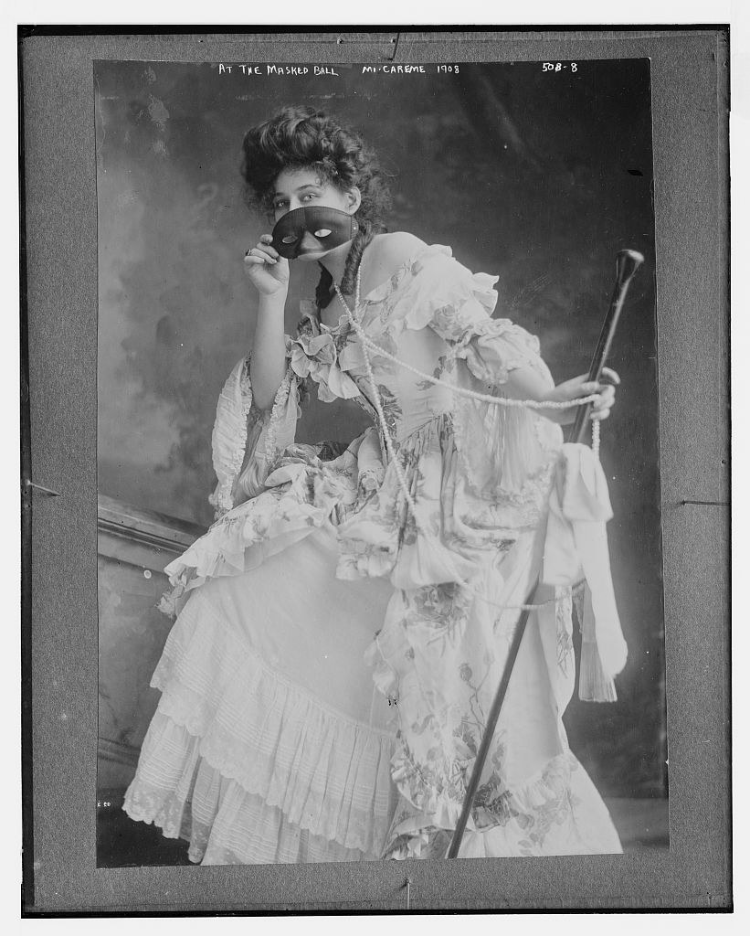 Woman in elaborate white Edwardian gown holding black masquerade mask, wearing long beads, posed for studio portrait, 1908.