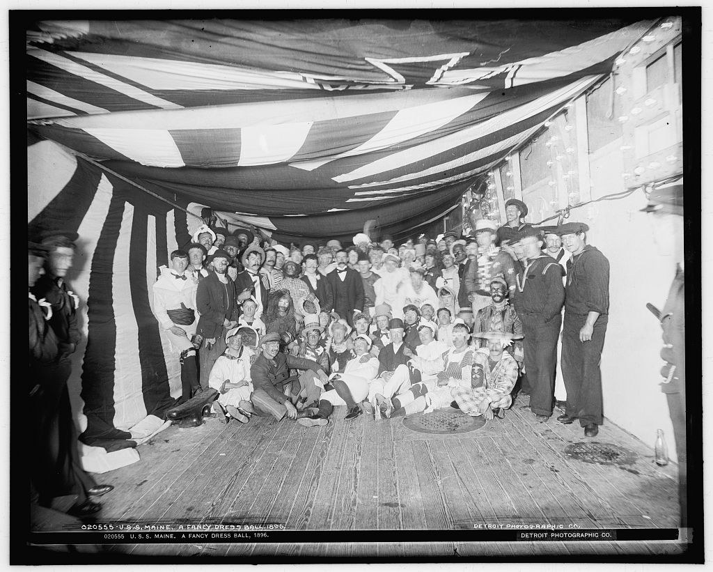 Crowded formal gathering of about 100 men, women, and children in 1890s dress aboard a ship, seated and standing beneath striped fabric canopy.