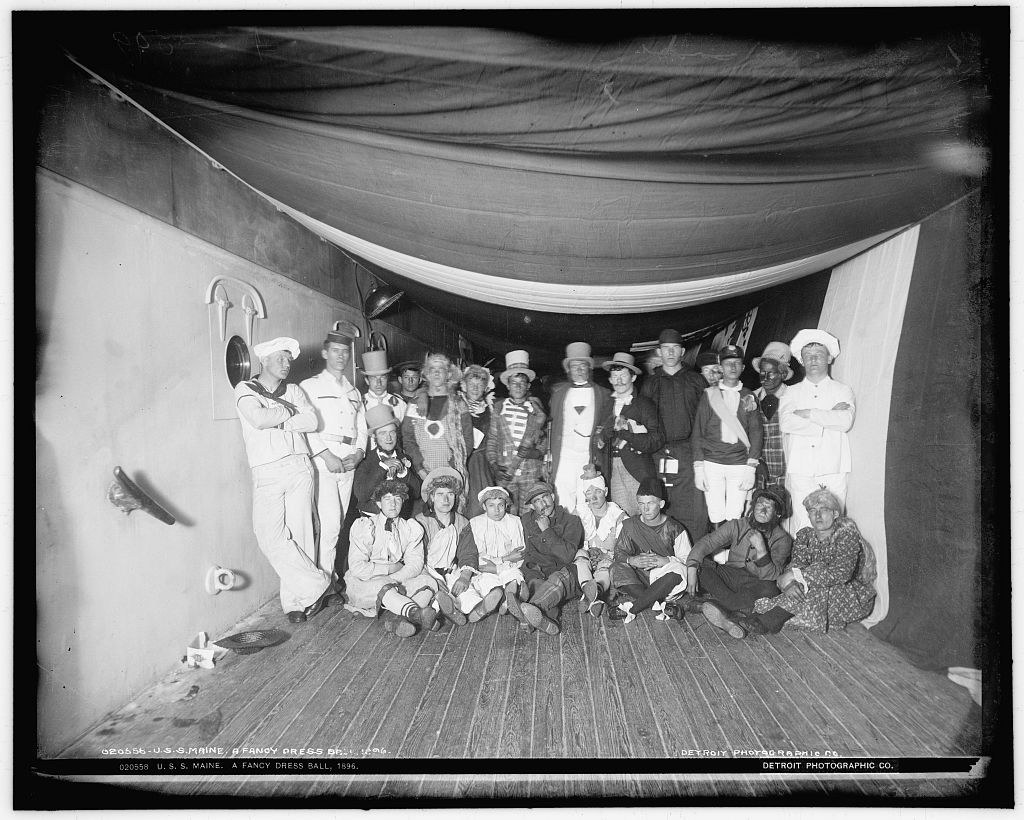 Thirty people in elaborate costumes and formal dress stand and sit in the wooden hold of a ship beneath draped fabric, posed for a group portrait.