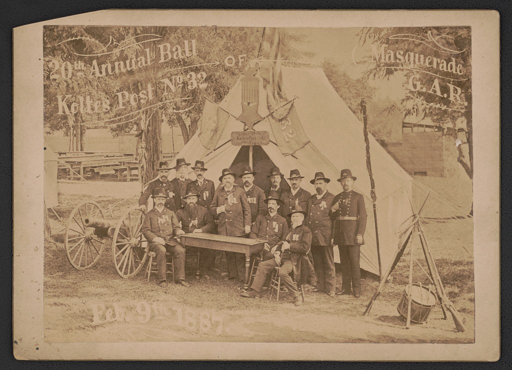 Sepia photograph of approximately twenty uniformed Civil War veterans in formal dress posed in and around a large canvas tent, February 1887.