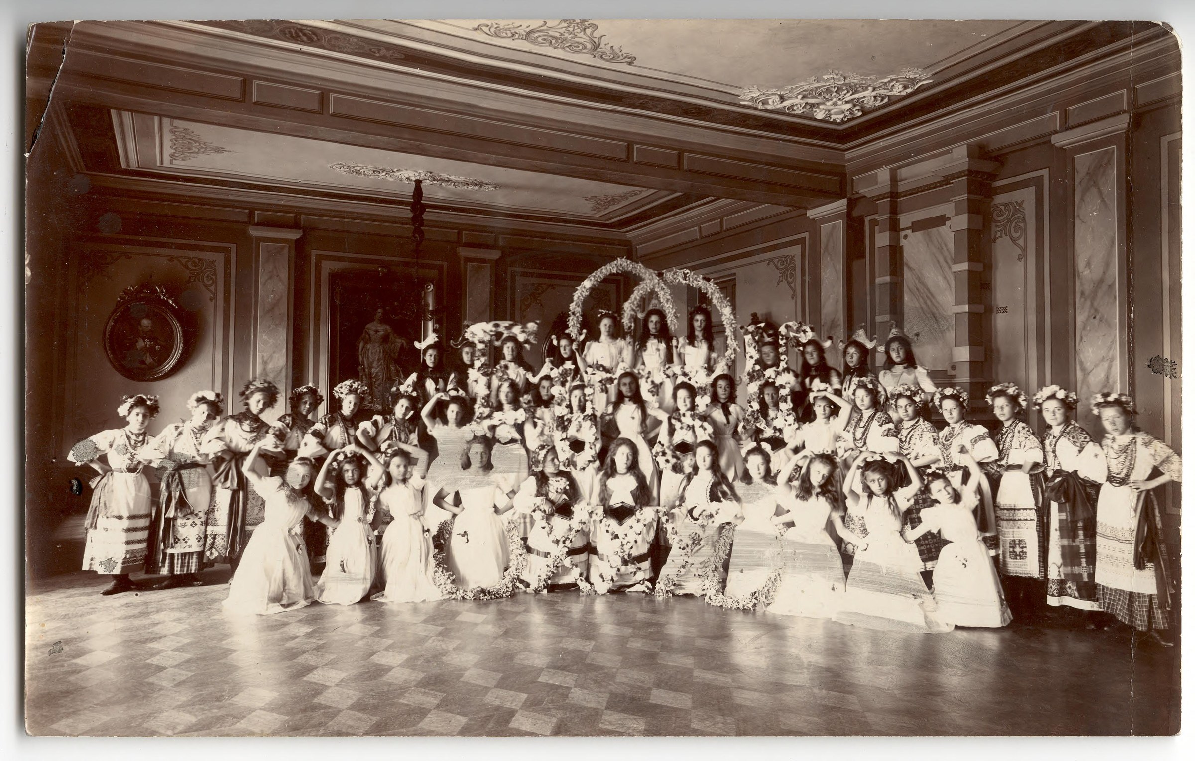 Formal group photograph of approximately sixty women and men in white evening dress and Russian national costume gathered in an ornate ballroom around a large