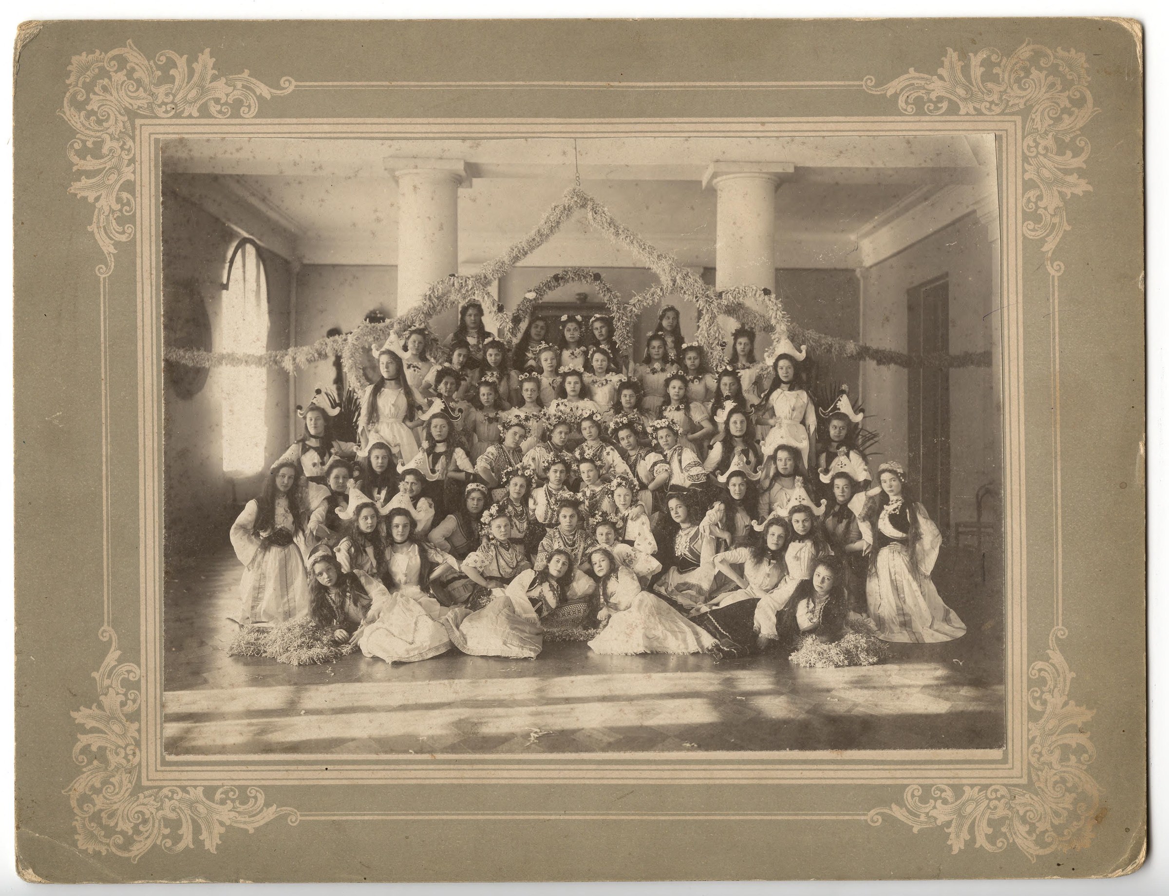 Over eighty young women in white gowns fill a grand hall around a central decorated pavilion at a formal ball, 1902.