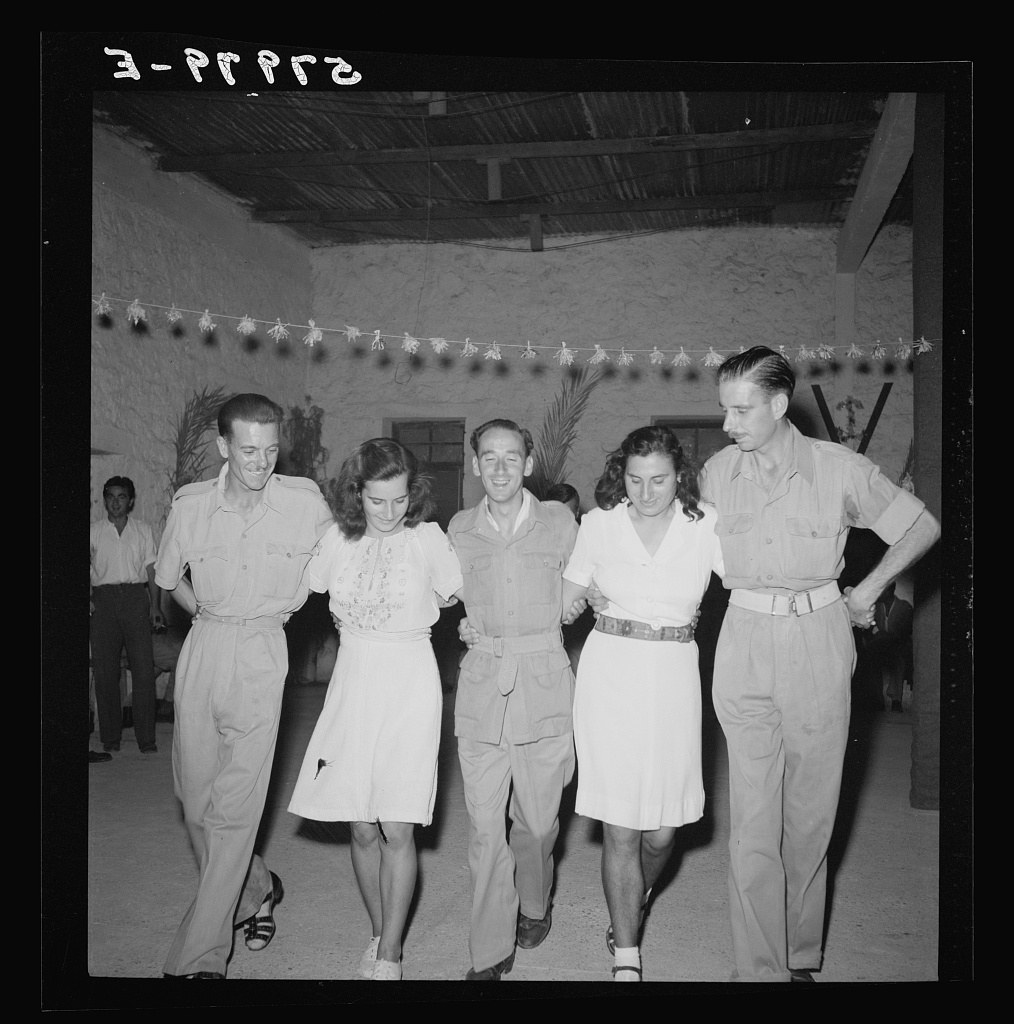 Five people in light-colored clothing stand in a line in a bare shed with a corrugated roof, posed facing the camera.