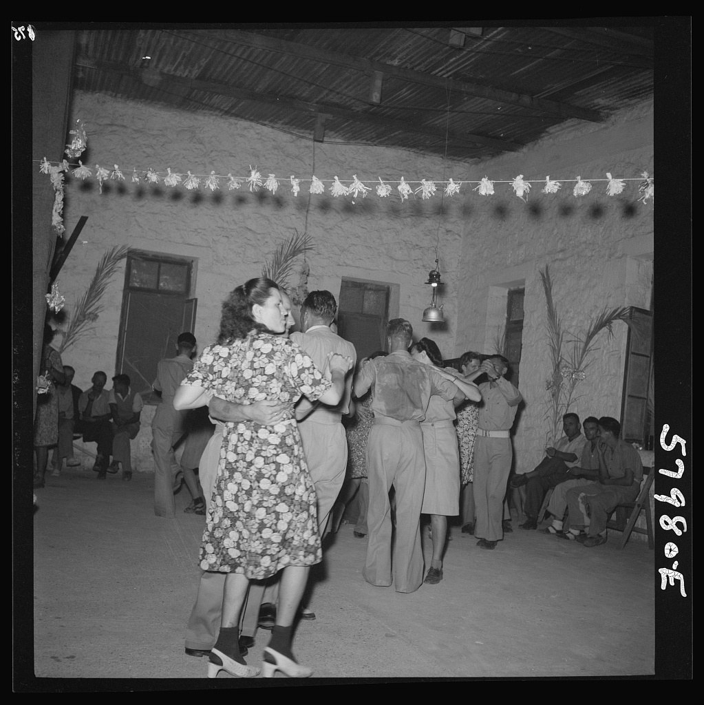 Couples dance in close embrace in a large hall decorated with paper cutouts; onlookers sit and stand around them in a refugee camp.