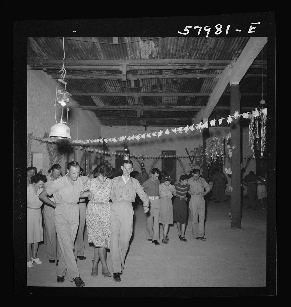 Approximately twenty people of mixed ages dance together in a metal-roofed shelter decorated with paper garlands, El Shatt refugee camp, 1944.