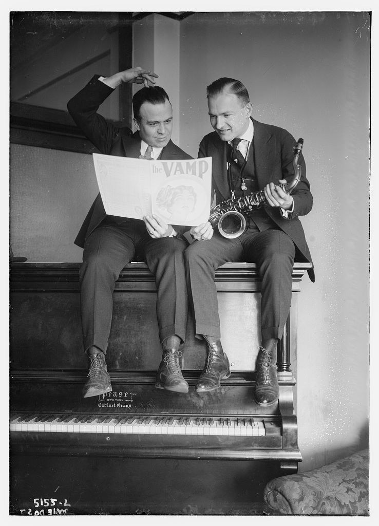 Two men in suits sit on a piano bench. The left man holds a sheet marked 'VAMP'; the right holds a saxophone.