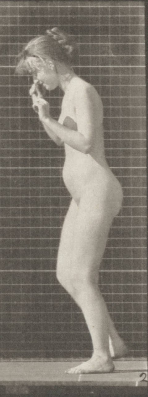 Barefoot unclothed woman in profile against brick wall, standing upright with weight distributed between two legs, arms at sides.