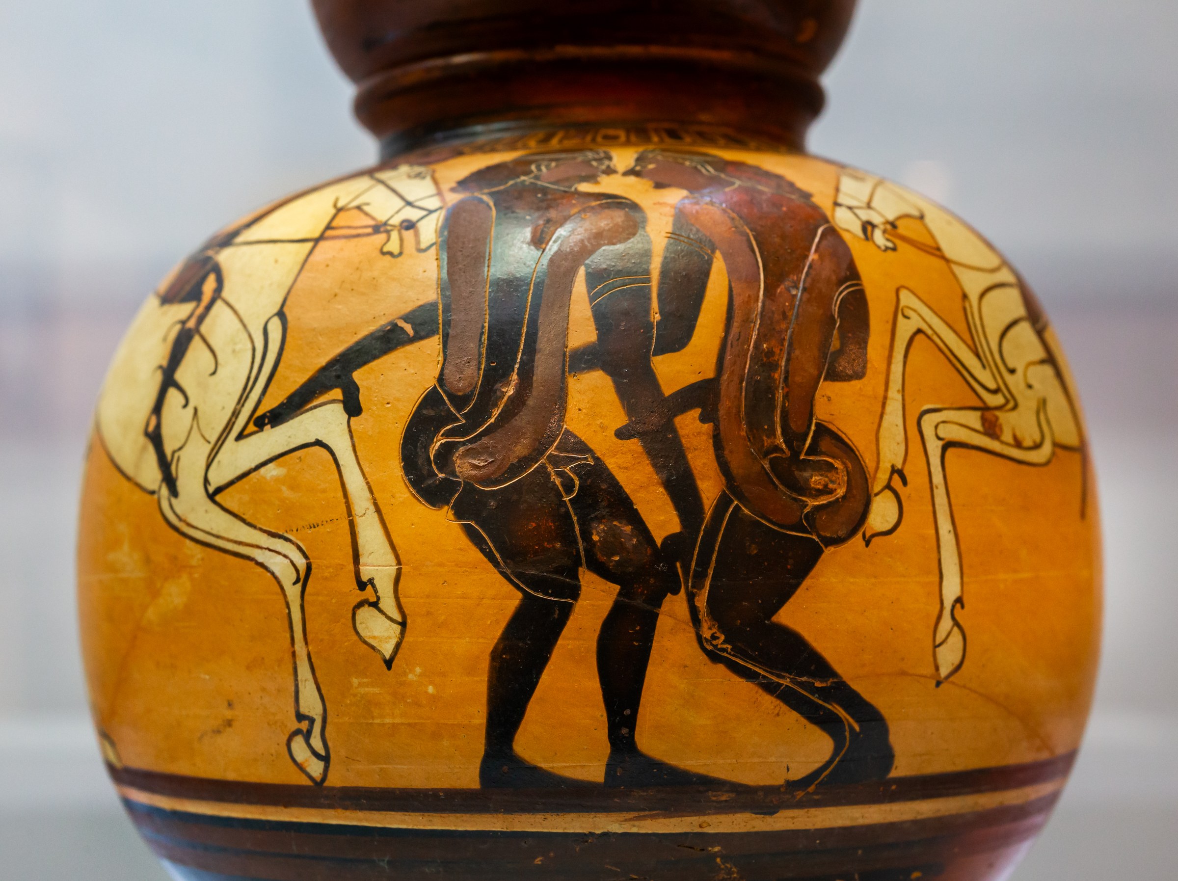 Two padded dancers in exaggerated forward-bent posture flank two horsemen on an ancient Greek pottery vessel.