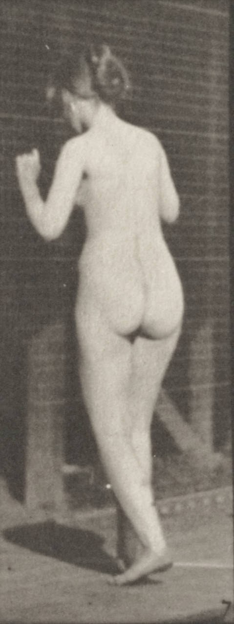 Nude woman photographed from behind in a contrapposto stance, one leg straight, one bent, arm extended, against a dark background.