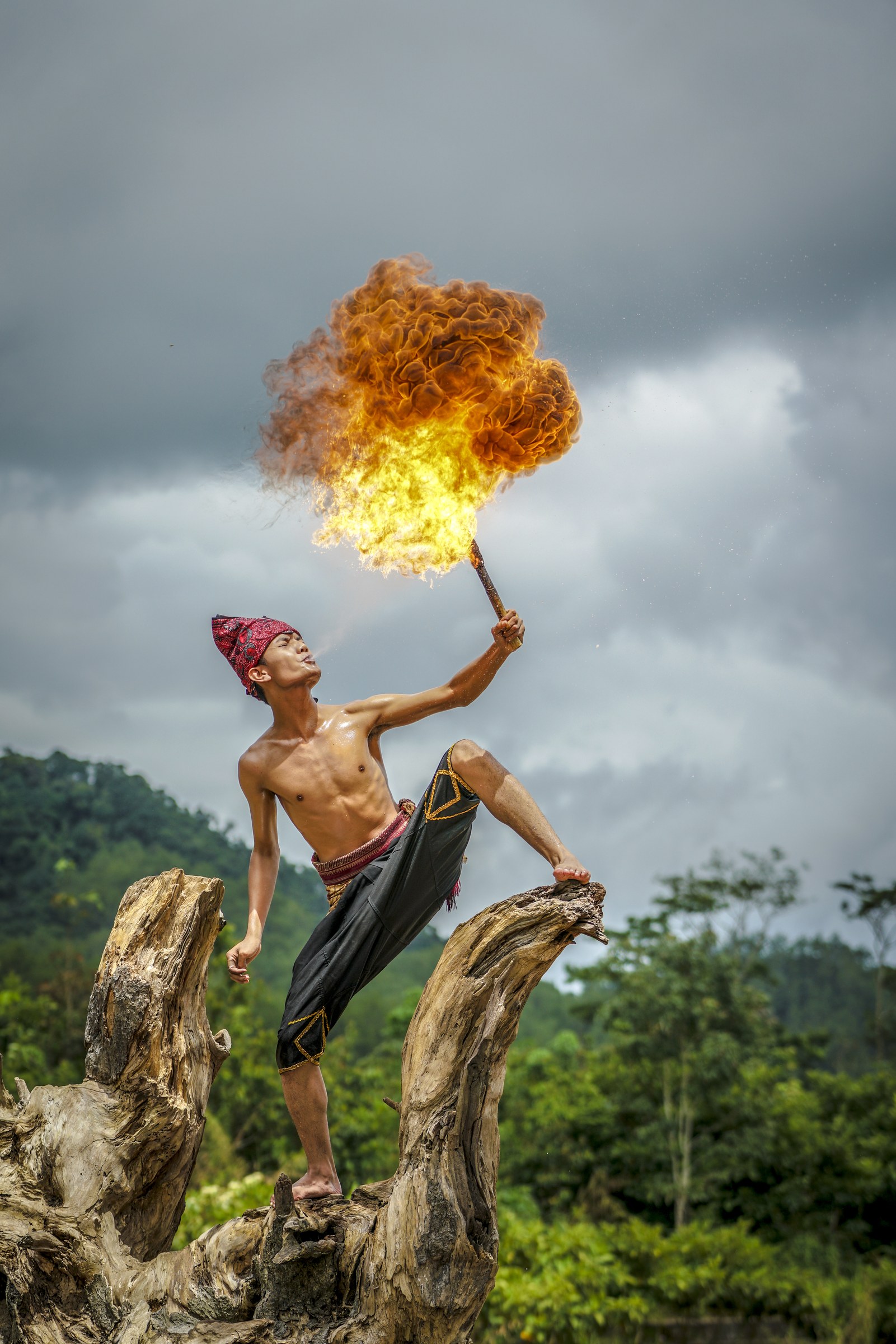 Shirtless male dancer in extreme backbend on tree stumps, holding flaming staff high overhead against grey sky and jungle backdrop.