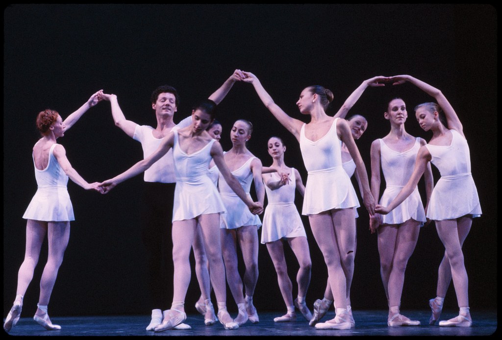 Ten dancers in pale tutus stand in a circle en pointe, arms linked and raised, against a black background.