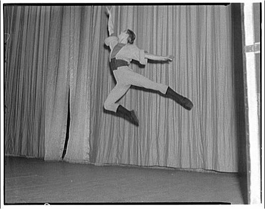Male ballet dancer captured mid-leap in a scissor split against a corrugated metal wall, shot from above in black and white.