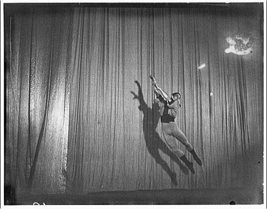 Black-and-white photograph of a classical ballet dancer in mid-leap, arms raised, wearing a tutu against a curtained backdrop, circa 1920.