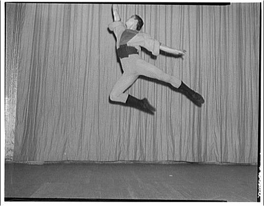 Male ballet dancer in mid-leap against corrugated metal backdrop, legs split vertically, arms spread wide.