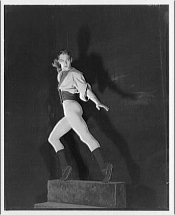 Black and white photograph of a female ballet dancer mid-leap, body arched, against a dark studio background.