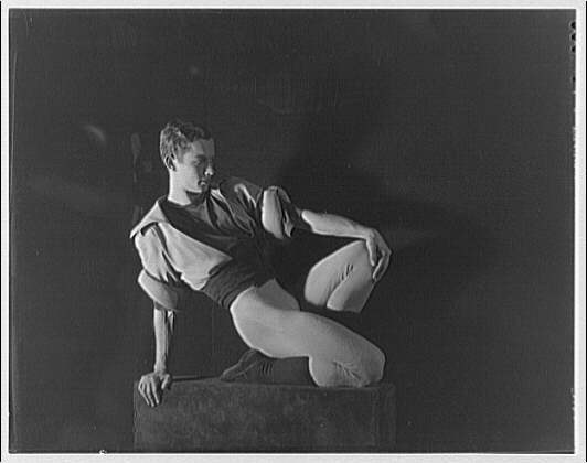 Black-and-white photograph of a solitary male ballet dancer seated on a platform, legs extended, arm supporting torso, in dramatic side-lighting against dark