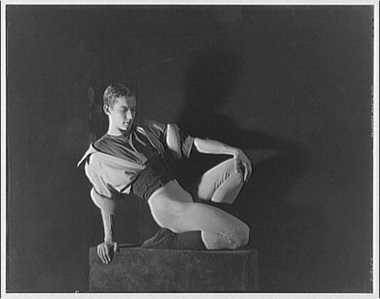 A male ballet dancer seated on a platform, posed in profile, wearing a dark tunic and tights against a dark background.