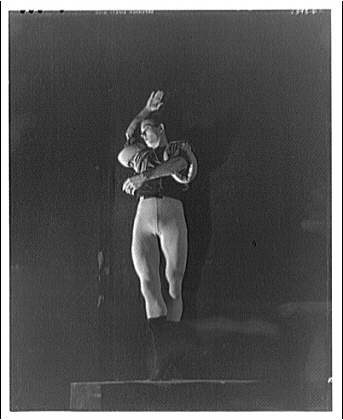 Black-and-white photograph of a female ballet dancer in a light tunic, standing in classical ballet position with one arm raised, against a dark background.