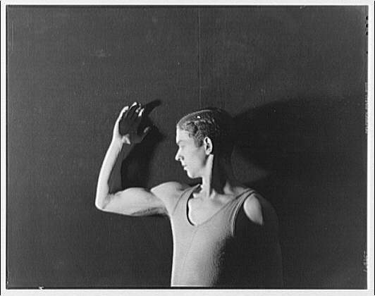 A shirtless male dancer stands in profile, right arm raised and bent near his head, against a dark background.