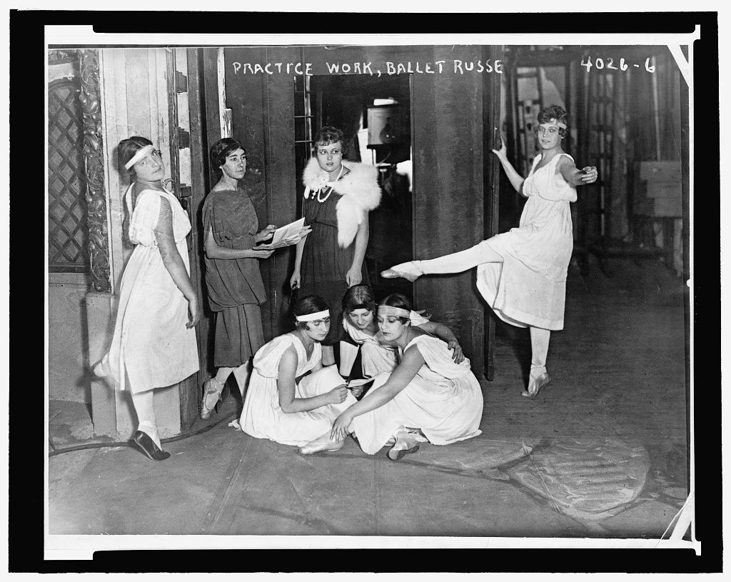 Seven young women in tunics and headbands during a ballet rehearsal, some standing in classical positions, others crouching on a bare studio floor.