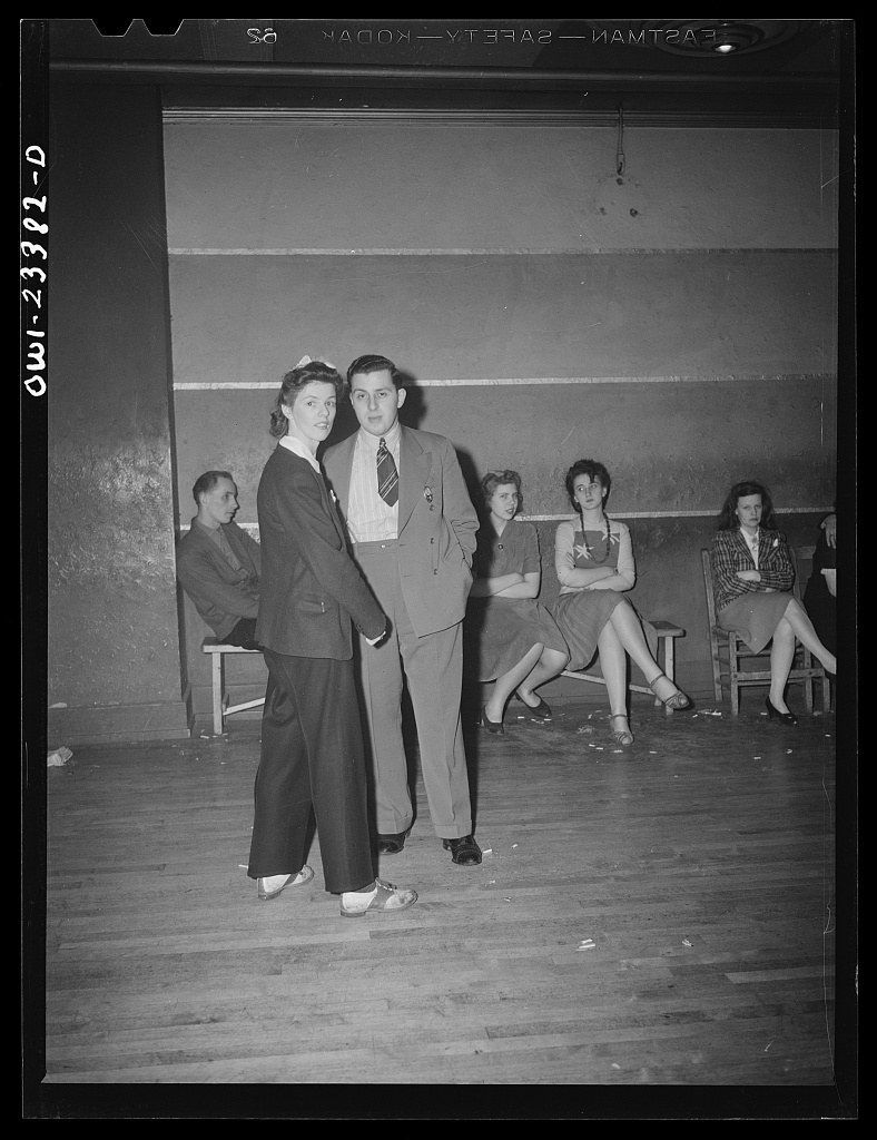 A man and woman dance swing in the center of a dance hall while five people watch from chairs in the background.