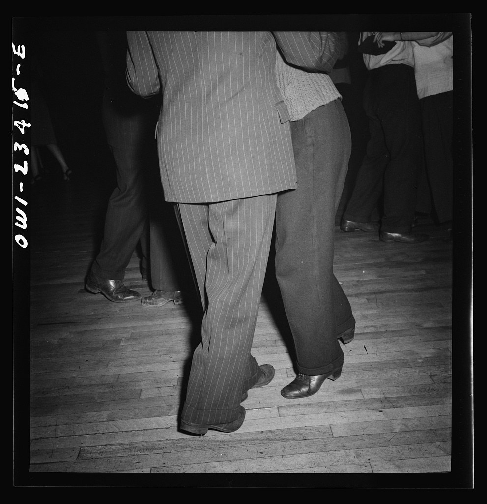 Two dancers in a crowded ballroom, photographed from the knees down. The man wears a light pinstriped suit; the woman wears dark stockings. Their feet show a