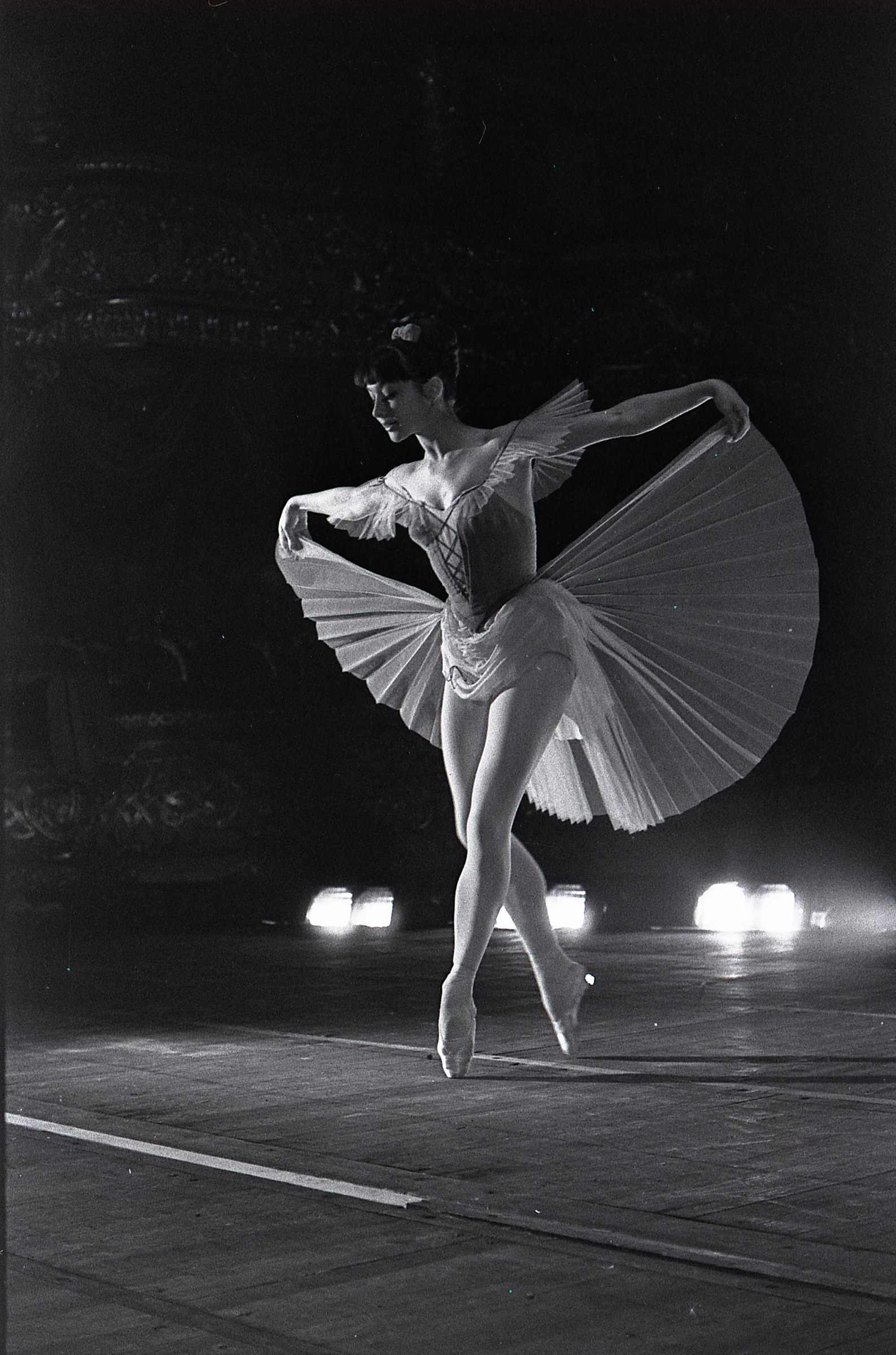 Female ballet dancer on pointe in white tutu, arabesque pose, against dark background with theatrical lighting.