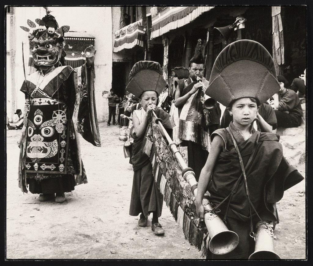 Tibetan Buddhist monks in ceremonial robes and headdresses process through a monastery courtyard, one wearing a large wrathful deity mask, others playing long