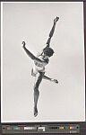 Male dancer in dark attire mid-leap against plain background, arms and one leg extended in balletic arabesque position.